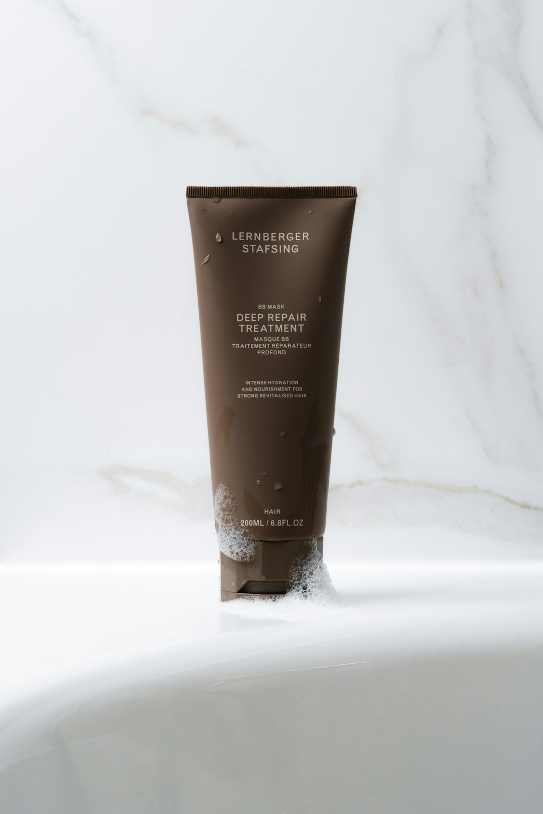 services-01 This image showcases a deep repair treatment in a sleek, matte brown tube with minimalist white text. The packaging is modern and sophisticated, complementing the clean aesthetic of the white marble background. The tube is slightly wet, with small soap bubbles surrounding the base, emphasizing its use as a hair care product. Positioned on the edge of a bathtub, the setting suggests a spa-like or self-care routine. The composition is balanced, with soft lighting enhancing the neutral tones and textures in the scene.
